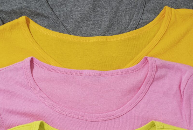 Image de 4 t-shirts de couleurs (gris, orange, rose et jaune) empilés.